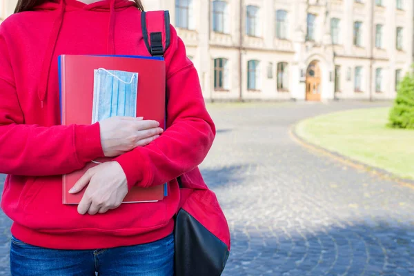 First day at school after university concept. Cropped close up photo of teen girl in casual red sweater holding books and medical mask standing near doors enter