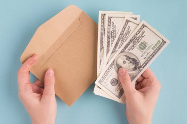 Yukarıdan bakan ilk kişi kadın ellerinin Amerikan dolarlarını tutarken çekilmiş fotoğrafı ve mavi renkli pastel arka planda izole edilmiş el işi kağıt zarf.