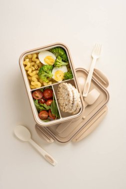 Brokoli, haşlanmış yumurta, makarna, pirinç keki ve domateslerden oluşan dikey hazırlanmış bir yemek, uygun ve besleyici bir yemek için yeniden kullanılabilir bir bölme kabında sunulmaktadır.