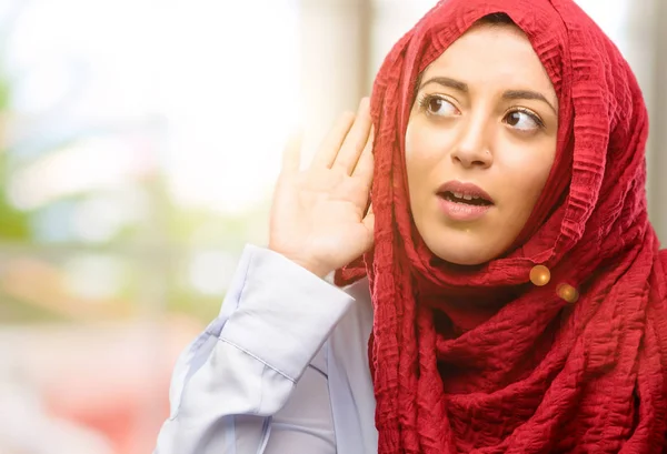 Young arab woman wearing hijab holding hand near ear trying to listen to interesting news expressing communication concept and gossip