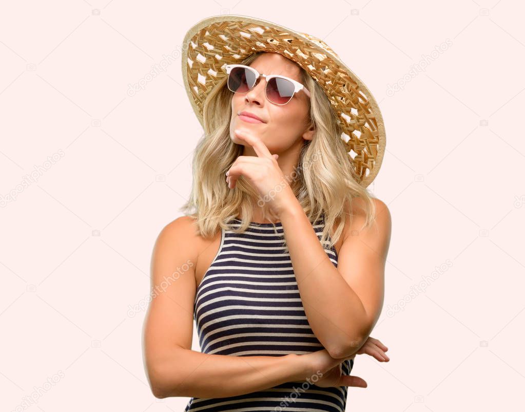 Young woman using sunglasses wearing summer hat thinking and looking up expressing doubt and wonder