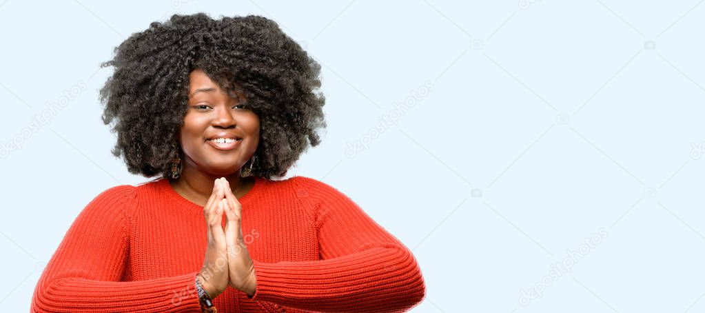 Beautiful african woman with hands together in praying gesture, expressing hope and please concept, blue background