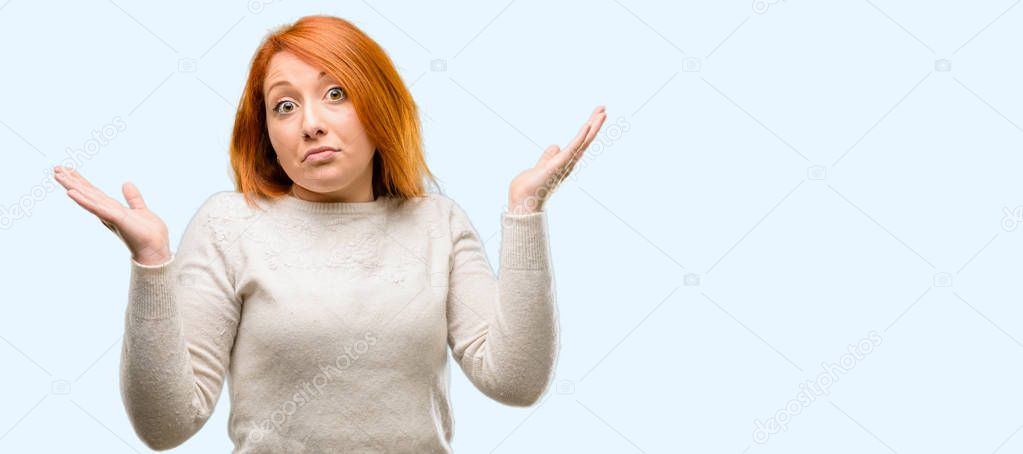 Beautiful young redhead woman doubt expression, confuse and wonder concept, uncertain future shrugging shoulders isolated over blue background