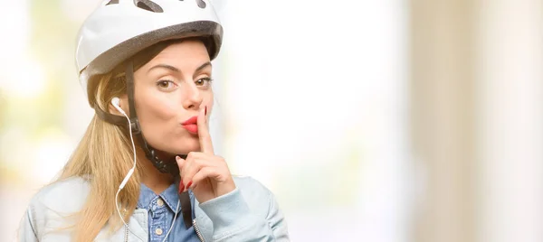 Young woman with bike helmet and earphones with index finger on lips, ask to be quiet. Silence and secret concept