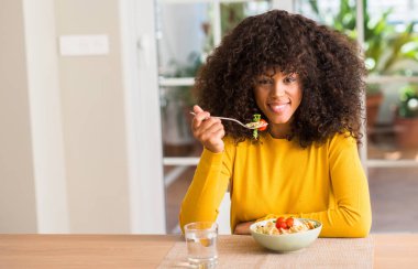 Afrikalı-Amerikalı kadın ayakta ve diş gösterilen kendine güvenen bir gülümsemeyle gülümseyerek mutlu bir yüz ile makarna salatası evde yemek