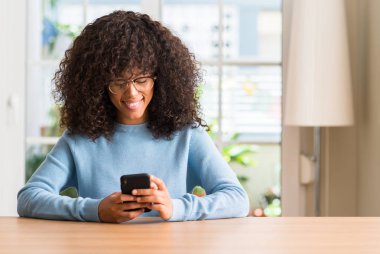 Güzel bir Afrikalı-Amerikalı kadın ayakta ve diş gösterilen kendine güvenen bir gülümsemeyle gülümseyerek mutlu bir yüz ile Smartphone kullanarak