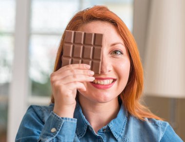 Evde duran ve diş gösterilen kendine güvenen bir gülümsemeyle gülümseyerek mutlu bir yüz ile çikolata bar tutan Kızıl saçlı kadın
