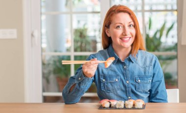 Kızıl saçlı kadın ayakta ve diş gösterilen kendine güvenen bir gülümsemeyle gülümseyerek mutlu bir yüz ile yemek çubukları kullanarak suşi yemek