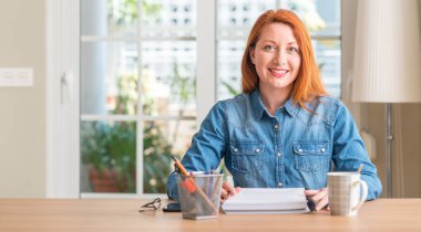 Kızıl saçlı kadın evde duran ve diş gösterilen kendine güvenen bir gülümsemeyle gülümseyerek mutlu bir yüz ile eğitim