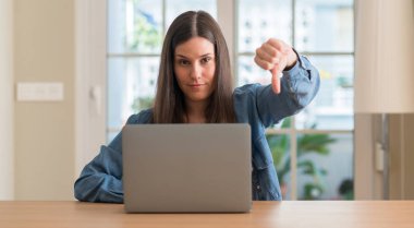 Dizüstü evde kullanan kızgın yüzü, eksi işareti aşağı antipati başparmak ile gösterilen, ret kavramı ile genç kadın