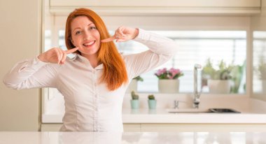 Kendinden emin gösterilen ve işaret parmakları diş ve ağız ile gülümseyen mutfak Kızıl saçlı kadın. Sağlık kavramı.