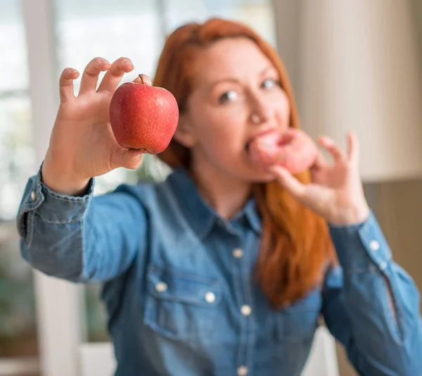 红头发的女人在苹果和甜甜圈之间选择一个快乐的脸站在微笑着自信的微笑显示牙齿 — 图库照片