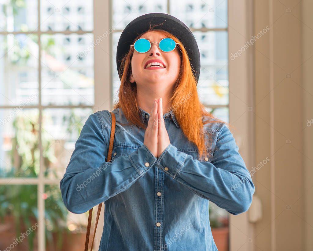 Stylish redhead woman wearing bowler hat and sunglasses begging and praying with hands together with hope expression on face very emotional and worried. Asking for forgiveness. Religion concept.