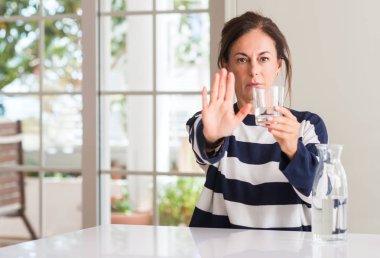 Orta yaşlı kadın açık eliyle dur işareti ile ciddi ve kendine güvenen ifade, savunma jest yapıyor bir bardak su içme
