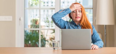Redhead woman using computer laptop at home stressed with hand on head, shocked with shame and surprise face, angry and frustrated. Fear and upset for mistake. clipart