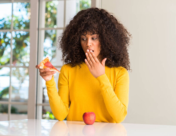 African american woman choosing between apple and pizza slice cover mouth with hand shocked with shame for mistake, expression of fear, scared in silence, secret concept