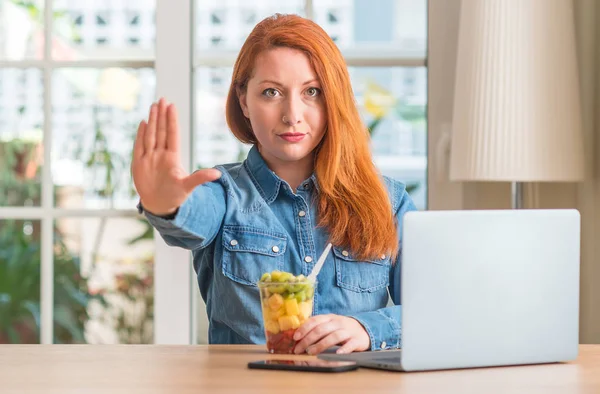 Redhead woman using computer laptop eating fruit at home with open hand doing stop sign with serious and confident expression, defense gesture