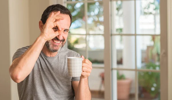 中年男子喝咖啡与快乐的脸微笑做 标志用手在眼睛看通过手指 — 图库照片