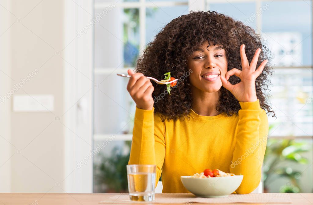 African american woman eating pasta salad at home doing ok sign with fingers, excellent symbol