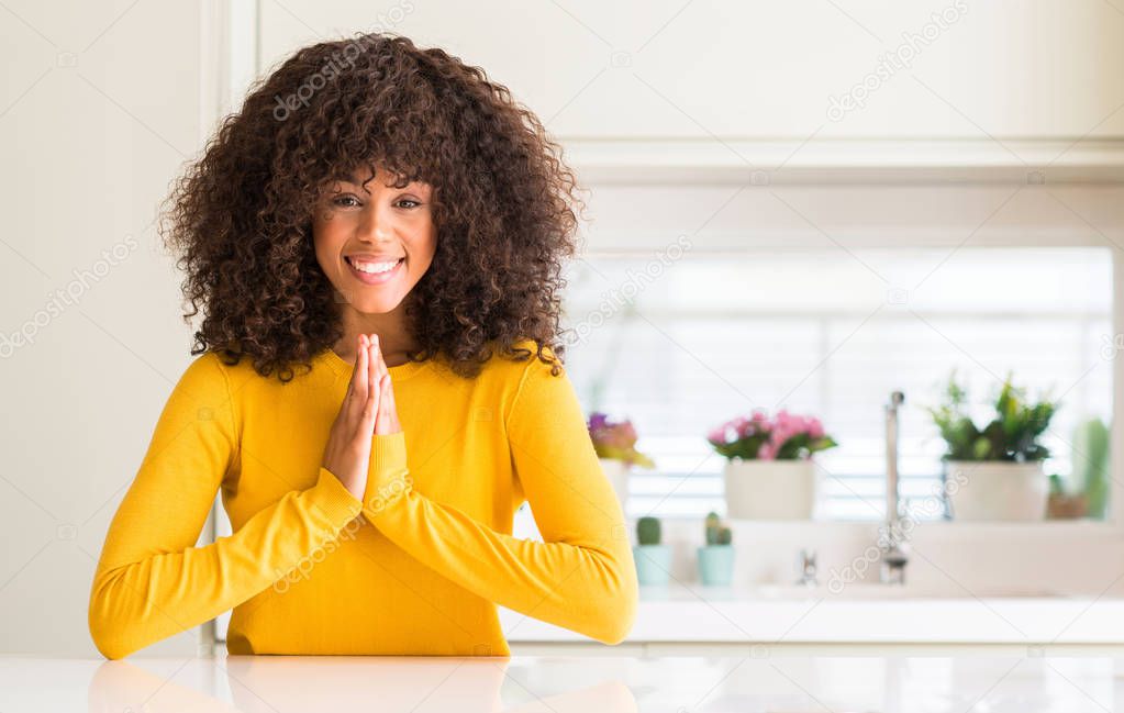 African american woman wearing yellow sweater at kitchen praying with hands together asking for forgiveness smiling confident.