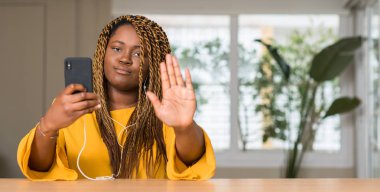 Afrikalı-Amerikalı kadın akıllı telefon istimal headphones ile açık eliyle dur işareti ile ciddi ve kendine güvenen ifade, savunma hareketi yapıyor