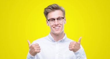 Genç yakışıklı sarışın adam başarı işareti başparmak gülümseyen ve mutlu el ile olumlu hareketi yapıyor. Neşeli bir ifadeyle, kazanan jest kameraya bakıyor.