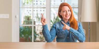 Kızıl saçlı kadın evde gülümseyen ve iki elleri ve parmakları ile tarafına işaret kamera bakıyor.