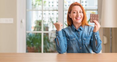 El ve parmak ile yukarı dönük mutlu yüz gülümseyen ile evde çikolata bar tutan Kızıl saçlı kadın