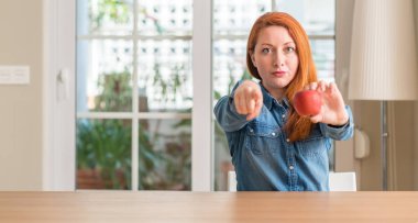 Kırmızı elma evde tutan Kızıl saçlı kadın kamera ve size, parmak ile işaret el işareti, pozitif ve kendinden emin bir jest ön