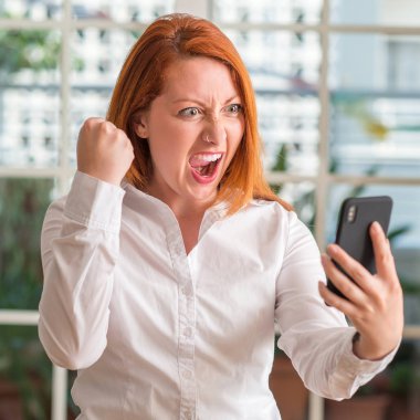 Akıllı telefon kullanan ev rahatsız ve hayal kırıklığı ile öfke, çılgın ve yükseltilmiş eliyle diye bağıran öfke kavramı bağırarak Kızıl saçlı kadın