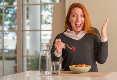 Kızıl saçlı kadın yeme meyve kase, kivi ve büyük gülümseme ile çığlık zaferini kutluyor çilek evde çok mutlu ve heyecanlı, kazanan ifade ve ellerini kaldırdı