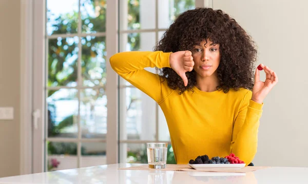 非洲裔美国妇女吃覆盆子和蓝莓在家中与愤怒的脸 消极的迹象显示不喜欢拇指下来 拒绝概念 — 图库照片