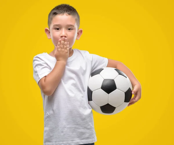 Dark haired little child playing with soccer ball cover mouth with hand shocked with shame for mistake, expression of fear, scared in silence, secret concept