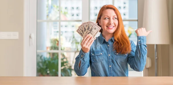 Рыжая Женщина Держащая Дома Банкноты Долларах Указывая Рукой Пальцем Вверх — стоковое фото