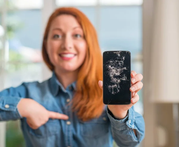 Redhead woman holding broken smartphone with surprise face pointing finger to himself