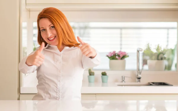红头发的妇女在厨房批准做积极的手势与手 竖起大拇指微笑 并为成功高兴 看着相机 胜利者的手势 — 图库照片