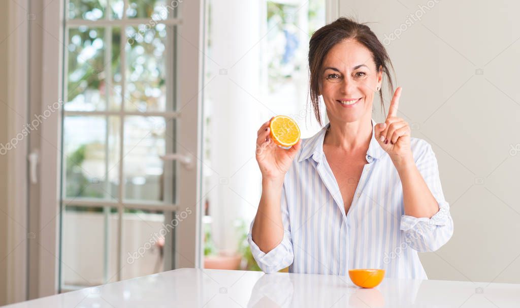 Middle aged woman holding orange fruit surprised with an idea or question pointing finger with happy face, number one