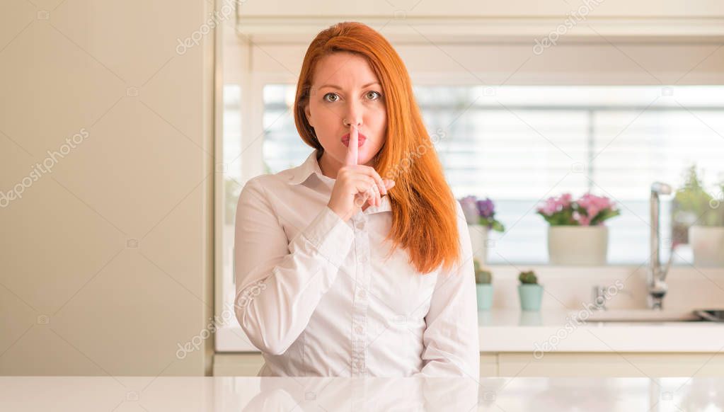 Redhead woman at kitchen asking to be quiet with finger on lips. Silence and secret concept.