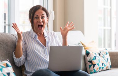 Kocaman bir gülümseme ile çığlık zaferini kutluyor ve ellerini kaldırdı kanepe çok mutlu ve heyecanlı, kazanan ifade içinde dizüstü bilgisayar kullanan kadın orta yaşlı