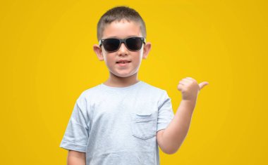 Koyu saçlı küçük çocuk el ve parmak ile yukarı dönük mutlu yüz ile gülümseyen güneş gözlüğü takıyor