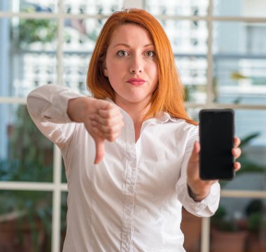 Evde Smartphone kullanan kızgın yüzü, eksi işareti aşağı antipati başparmak ile gösterilen, ret kavramı ile Kızıl saçlı kadın