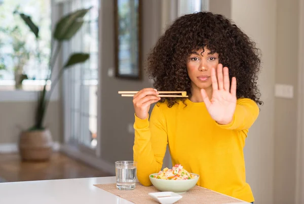 African american woman eating asian rice at home with open hand doing stop sign with serious and confident expression, defense gesture