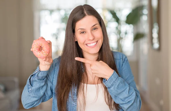 拿着粉红色甜甜圈的饥饿的年轻妇女用手和手指非常愉快地指向 — 图库照片