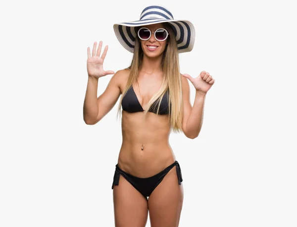 Beautiful young woman wearing bikini, sunglasses and hat showing and pointing up with fingers number six while smiling confident and happy.