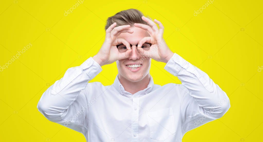 Young handsome blond man doing ok gesture like binoculars sticking tongue out, eyes looking through fingers. Crazy expression.