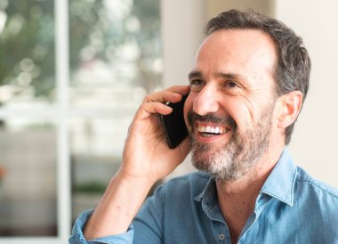 Ayakta ve diş gösterilen kendine güvenen bir gülümsemeyle gülümseyerek mutlu bir yüz ile akıllı telefon kullanan orta yaş adam