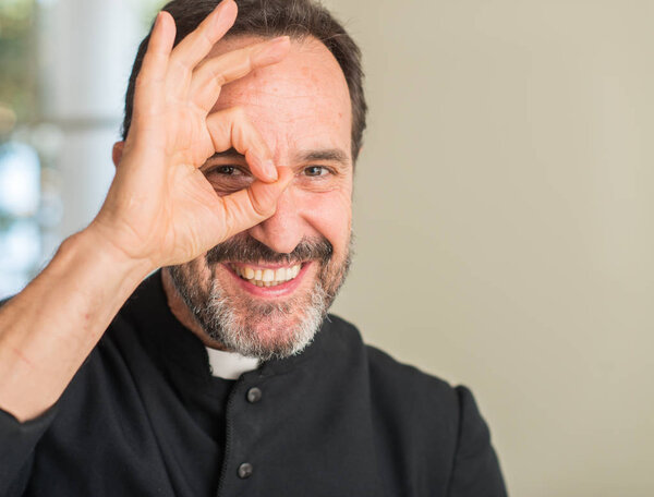 Christian priest man with happy face smiling doing ok sign with hand on eye looking through fingers
