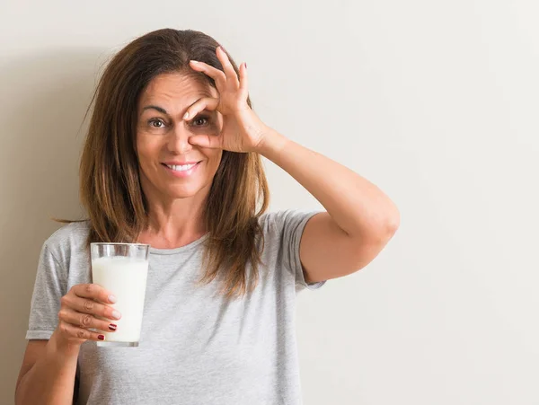 中年妇女喝一杯新鲜的牛奶与愉快的面孔微笑做 标志用手在眼睛看通过手指 — 图库照片
