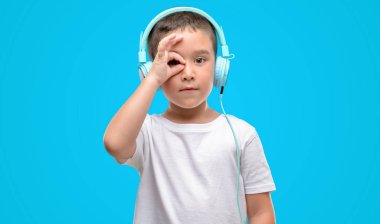 Koyu saçlı küçük çocuk müzik dinleme el parmaklarının arasından seyir göz üzerinde ok işaretiyle yapıyor gülümseyen mutlu yüz ile kulaklıklar ile