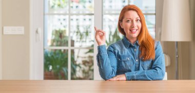 Kameraya bakarak yan el ve parmak ile işaret eden Kızıl saçlı kadın evde yüzünde kocaman bir gülümseme ile.